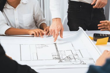 Mimari proje üzerinde birlikte çalışan çeşitli inşaat mühendisi ve müvekkil grupları, inşaat planını gözden geçirip toplantı masasında plan oluşturuyorlar. Tedbirli