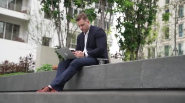 Yetenekli erkek lider Green City 'deki yatırım verilerini analiz ederken aynı zamanda dizüstü bilgisayarını kullanarak telefon gönderme raporu üzerinde çalışıyor. Parkta ya da şehir merkezinde otururken yatırımcı görünümlü mutlu pazarlama planı. Urbane.