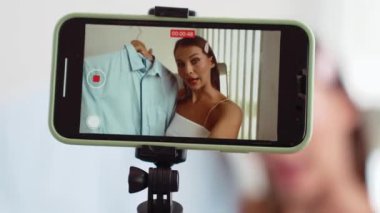 Genç sosyal medyum kadın moda videosu yapıyor. Blogcu kameraya selfie çubuğu ve ışık halkası kullanarak gülümserken, aynı zamanda ikna edici çevrimiçi kıyafetlerin izleyicilere veya takipçilere vlog satmasını sağlıyor. Düzenle