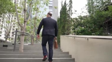 Yeşil şehirde bavulunu tutarken resmi takım elbiseli beyaz bir müdürün merdivenlerden çıkışının arka görüntüsü. Profesyonel bir iş adamı iş yerine giderken aynı zamanda Eko Şehri 'nde merdivenleri çıkıyor. Urbane..