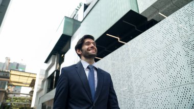 Takım elbiseli ve gülümseyen zeki bir iş adamı beyaz modern mimari binasının yanında sokakta yürürken. Şehir merkezinde çekici, yakışıklı bir yönetici gökdelen gibi görünüyor. Uzayı kopyala Sevinçli..