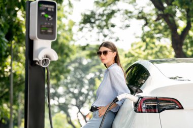 Yolculuk sırasında doğal orman ya da milli parktaki elektrikli araba için şarj olan genç bir kadın. Tatil ve tatil boyunca çevre dostu seyahat. Yükselt