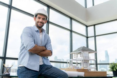 Güvenlik kaskı, proje planı ve ev modeli ile kameraya bakan profesyonel mimar mühendisinin portresi kendine güvenle poz verirken toplantı masasında duruyor. İnşaat mühendisliği, erkek işçi. İzleme.