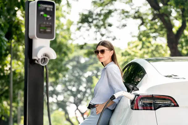 Yolculuk sırasında doğal orman ya da milli parktaki elektrikli araba için şarj olan genç bir kadın. Tatil ve tatil boyunca çevre dostu seyahat. Yükselt