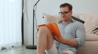 Orta yaşlı bir adam elinde kahve fincanıyla kanepede oturup ders kitabı okuyarak sosyal ve teknolojik meslekler hakkında bilgi artışı yapar ya da çocukları iyi bir yetişkin olarak yetiştirir. Hayat boyu öğrenme konsepti. Uyumsuzluk.