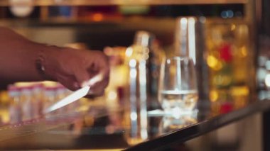 Kapanış barmeninin bardak altlığı üzerine şeffaf bardakta buz koyması, müşteriye servis yapmaya hazırlanırken arka planda içki tezgahı lüks barında el yapımı içecek içmesi. Vinosity.