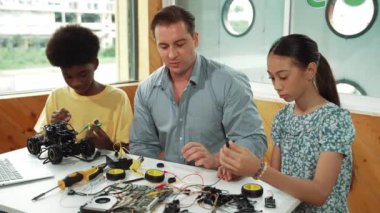 Akıllı akıl hocası araba modelini tamir ederken elektronik aletleri öğretiyor. Kafkas öğretmeni masadaki dizüstü bilgisayarlı robot inşaatı açıklarken, araba modeli etrafa saçılmış. Öğretim