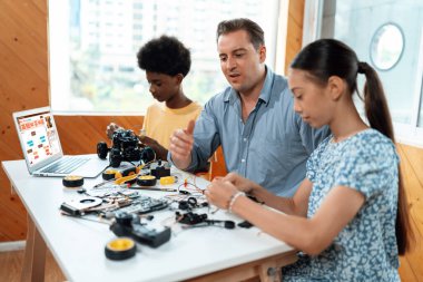Öğretmen öğrencilere robotik araba kodlamasını öğretirken aynı zamanda BTMM sınıfında mühendislik öğreniyor. Masaya dizüstü bilgisayar, kumanda, elektrik kablosu, şarj aleti, robot araç. Öğretim.