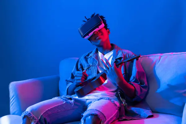 stock image Young African American singer using through VR glasses enjoy singing song with guitar playing on meta blue neon light wall virtual reality metaverse fantasy technology sitting on sofa. Contrivance.