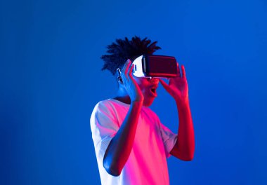 Sanal gerçekliği dijital fütüristik teknolojiye bağlayan pembe mavi neon duvarda VR 'dan bakan genç Afrikalı Amerikalı' ya şaşırdım. Entrika.