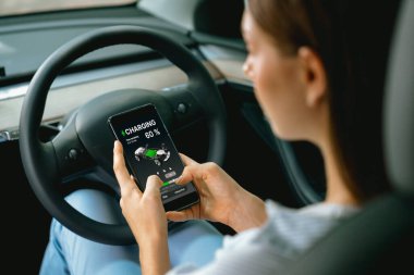 Aracın içindeki genç kadın, modern EV arabasındaki akıllı telefon ekranında akü şarj etme uygulamasını kontrol ediyor. Yükselt