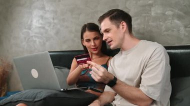 Genç çift, kredi kartıyla ödeme yapmak için internet üzerinden ödeme uygulaması ve dijital cüzdan kullanarak yatak odasında oturuyor. Mobil internet üzerinden alışveriş ve modern alımlar. Düzenle