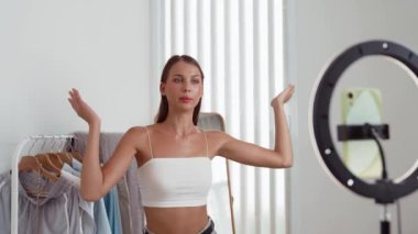 Genç sosyal medyum kadın moda videosu yapıyor. Blogcu kameraya selfie çubuğu ve ışık halkası kullanarak gülümserken, aynı zamanda ikna edici çevrimiçi kıyafetlerin izleyicilere veya takipçilere vlog satmasını sağlıyor. Düzenle