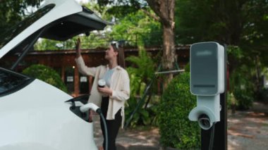 Yavaş çekimde çalışan kadın ve EV elektrikli araba ile sürdürülebilir şehir ulaşımı ilkbahar bahçesindeki açık kafede şarj oluyor, yeşil şehir sürdürülebilirliği ve çevre dostu EV otomobil. Yararlı