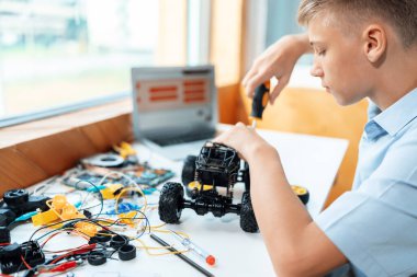 Sarı saçlı okul çocuğu, BTMM sınıfında robotik araç tamir eden bir tornavida kullanıyor. Masaya dizüstü bilgisayar, kumanda, elektrik kablosu, şarj aleti ve robot araç koyun. Öğretim.
