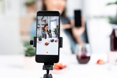 Seçici odaklı akıllı telefon ve gülümseyen genç, güzel Asyalı karışık çilek ve kiraz suyu detoksu değerlendirmesi sosyal medyada sağlıklı ürün tanıtımı sunuyor. Taktik.