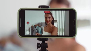 Genç sosyal medyum kadın moda videosu yapıyor. Blogcu kameraya selfie çubuğu ve ışık halkası kullanarak gülümserken, aynı zamanda ikna edici çevrimiçi kıyafetlerin izleyicilere veya takipçilere vlog satmasını sağlıyor. Düzenle