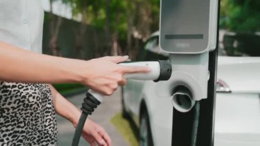 Şehir merkezindeki EV şarj istasyonunda elektrikli araba aküsünü yavaş çekimde şarj eden kadın elektrikli aracın iç organları için sürdürülebilir yeşil ve yenilenebilir temiz enerji yaşam tarzına sahip.