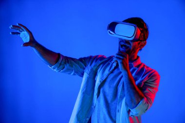 Sanal programı keşfetmek ve girmek için VR gözlük takan heyecanlı bir adam. Yetenekli oyuncular metaevrene dokunurken gündelik giysiler giyer ve neon ışıklı arka planda dururlar. Yaşam tarzı. Sapma.