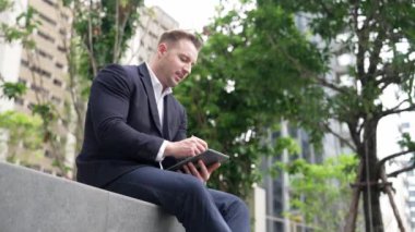 Tablet kalemle yazarken parkta oturan işadamının yan görüntüsü. Akıllı proje yöneticisinin finansal planı düşünürken veri analizine baktığı düşük açılı kamera. Urbane