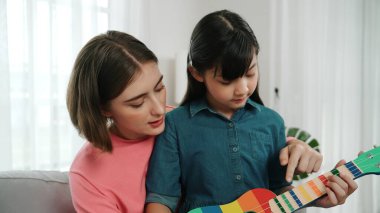 Kafkasyalı bir anne evde akustik müzik öğretirken ukulele çalan mutlu kız. Sevimli çocuk enstrüman öğreniyor. Kafkasyalı mutlu anne ve kız birlikte vakit geçiriyorlar. Pedagoji.