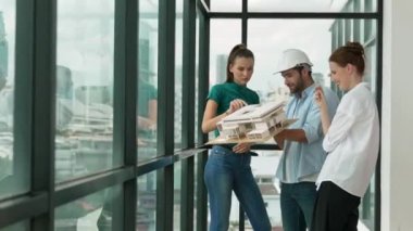 Proje yöneticisi ev modelini elinde tutarken, mimar mühendis takımı da ev tasarımı veya bina inşaatı hakkında fikir alışverişinde bulunuyor. Şehrin penceresinin yanında iç mimar duruyor. İzleme