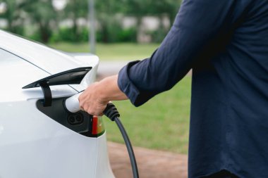 Açık yeşil şehir parkı manzaralı EV şarj istasyonundan EV elektrikli araçları şarj eden adam. Çevre dostu şehir ulaşımı ve çevre dostu EV araba seyahati. Yükselt