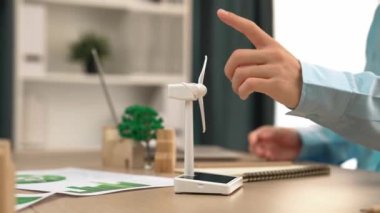 Temiz enerji ve ahşap blok kullanılarak temsil edilen yel değirmeni modelini izleyen iş adamı, gelecekteki yönetim için dizüstü bilgisayar ve defterle çalışan, eko ev dağınıklığını sembolize ediyor. Tanımlama.