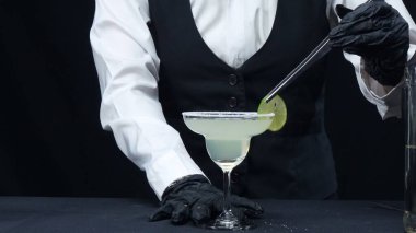 Profesyonel barmen taze margaritayı tuzlu garnitür bardağıyla süslüyor. Yetenekli barmen ellerinin makrografisi barda siyah arkaplanlı kokteyl yapmaya hazırlanıyor. Ağır çekim. Geliştirilebilir.