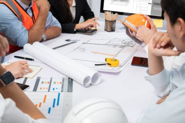 Mimari proje üzerinde birlikte çalışan çeşitli inşaat mühendisi ve müvekkil grupları, inşaat planını gözden geçirip toplantı masasında plan oluşturuyorlar. Tedbirli