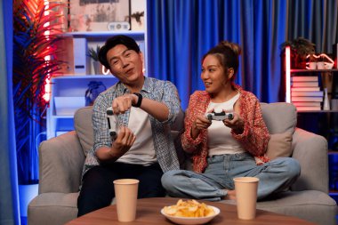 Neşeli, gülümseyen bir çiftin koltuğunda oturup oyun oynuyorlar. Oyun konsolu, hazır aperatifler ve içecekler, modern neon ışıklı, rahat oturma odasında eğlenceli bir rekabet seviyesinde. İnfobahn.