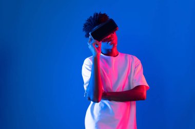 Sanal gerçekliği dijital fütüristik teknolojiye bağlayan pembe mavi neon duvarda VR 'dan bakan genç Afrikalı Amerikalı' ya şaşırdım. Entrika.