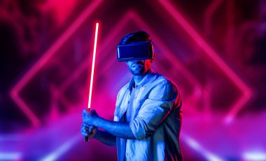 Lazer kılıcı tutan ve VR gözlük takan oyuncu. Beyaz adam, neon şatonun etrafında dikilirken görsel gerçeklik kulaklığı kullanıyor. Yenilik teknolojisi. Sapma.