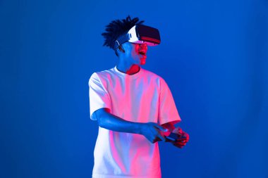 Genç Afrikalı Amerikalı oyuncu sanal gerçeklik dünyasını dijital fütüristik teknolojiye bağlayan mavi neon ışıklı duvarda akıllı telefon oyunu hologramı kullanarak VR kullanıyor. Entrika.