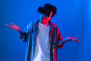 VR kullanan genç Afro-Amerikalı, boş uzay ürünlerini işaret ederek metaevren dünyasındaki mavi, neon ışıklı duvar fütüristik teknolojisi hakkında detaylı bilgi veriyor. Entrika.