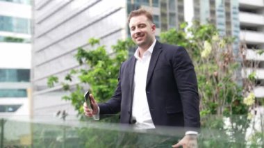 Beyaz yetenekli bir iş adamı, çatıda kameraya bakıyor. Yeşil şehirdeki modern binada resmi takım elbise giyerken kendine güvenen profesyonel bir yatırımcı. Yaşam tarzı. Urbane