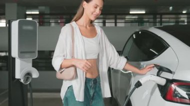 Yavaş hareket eden kadın mutlu ve EV elektrikli arabasıyla şehir merkezindeki Shopp Center otoparkına doğru gülümsüyor. Elektrikli taşıt iç organları için EV şarj prizi taşıyor.
