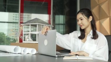 Genç ve güzel mimar mühendisin çatı malzemelerini kontrol etmesi ve ofisteki masaya yerleştirilmiş proje planı ve ev modeliyle dizüstü bilgisayarı kullanması. Yaratıcı iş tasarımı konsepti. Yönlendirici.