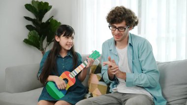 Akıllı kız oturma odasında ukulele çalarken beyaz bir babanın motivasyona ve teşviğe el çırpması. Amerikalı kız babasına gösterirken gitar öğreniyor. Aile yaratma kavramı. Pedagoji..