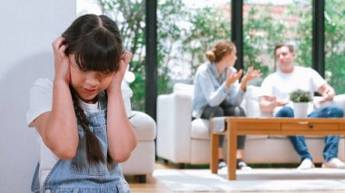 Stresli ve mutsuz genç kız köşeye kıvrılıp kulaklarını kapatarak arka planda ebeveyninin tartışmasını engelliyor. Evde aile içi şiddet ve travmatik çocukluk depresyona dönüşür. Eşzamanlama