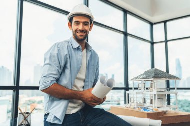 Güvenlik kaskı, proje planı ve ev modeli ile kameraya bakan profesyonel mimar mühendisinin portresi kendine güvenle poz verirken toplantı masasında duruyor. İnşaat mühendisliği, erkek işçi. İzleme.