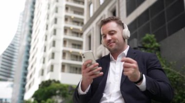 Kafkasyalı iş adamı cep telefonunu tutarken rahatlatıcı müzik dinliyor. Yetenekli mutlu yönetici müzik açar ve yeşil şehir veya parkta otururken neşeli bir şekilde şarkıya geçer. Urbane.