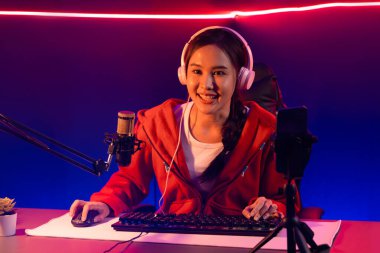 İnternet üzerinden kulaklık takıp gülümseyen Asyalı güzel kız flamasının sunucu kanalı izleyicilerle internet üzerinden konuşma yapıyor. Esport yetenekli takım oyuncuları neon mavi odada. Taktik.