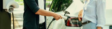 Odaklanmış EV arabanın pili, tatilde ulusal park ve yeşillik ormanlarında çevre dostu elektrikli arabalarla seyahat eden sevgili çiftin bulanık arka planına şarj oluyor. Yükselt