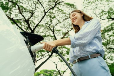 Yolculuk sırasında doğal orman ya da milli parktaki elektrikli araba için şarj olan genç bir kadın. Tatil ve tatil boyunca çevre dostu seyahat. Yükselt