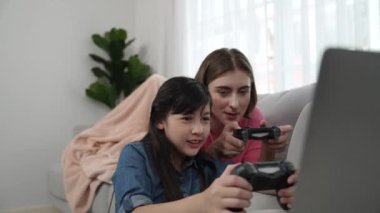 Annem dizüstü bilgisayar kullanarak kanepeye uzanırken, mutlu kız oyun oynuyor ve oturuyor. Beyaz bir ebeveyn ve çekici bir kız, elinde joystick ile birlikte vakit geçirir ve maç kazanmaya odaklanırlar. Pedagoji.