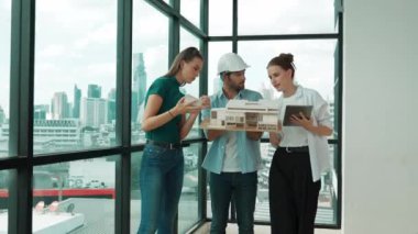 Proje yöneticisi ev modelini elinde tutarken, mimar mühendis takımı da ev tasarımı veya bina inşaatı hakkında fikir alışverişinde bulunuyor. Şehrin penceresinin yanında iç mimar duruyor. İzleme