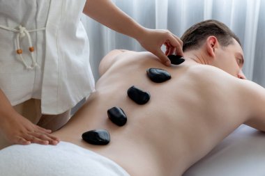 Lüks tatil köyündeki kaplıcada sıcak taş masajı. Gün ışığı dinginliği ortamı, mutlu erkek müşteri bazalt taşı masajının keyfini çıkarıyor. Rahatlatıcı bir sıcaklıkla vücudun üzerinde süzülüyor. Sessiz.