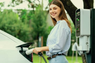 Açık yeşil şehir parkı manzaralı EV şarj istasyonundan genç kadın EV elektrikli araçları şarj ediyor. Çevre dostu şehir ulaşımı ve çevre dostu EV araba seyahati. Yükselt