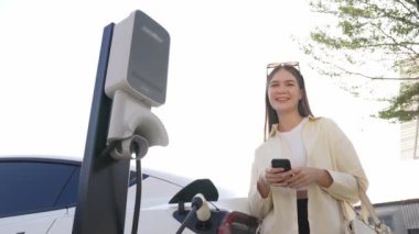 Ağır çekimde genç bir kadın, alışveriş merkezinin otoparkındaki şarj istasyonundan şarj olan EV 'nin pilini şarj ederken akıllı telefondan akü durumunu kontrol ediyor. Modern kadın elektrikli araçla alışverişe gider. Yararlı
